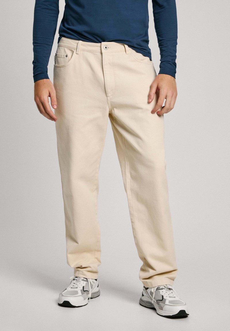 Pantalones de algodón beige con un corte recto, que cuentan con bolsillos frontales y cierre de botón, combinados con zapatillas deportivas en gris y blanco.