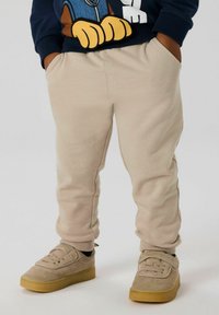 Kind trägt beige Sweatpants und hellbraune Turnschuhe, Hände in den Taschen, dazu einen marineblauen Pullover mit den großen gelben Pfoten einer Cartoonfigur.