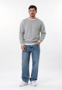 Άντρας που στέκεται με το πρόσωπο προς τα εμπρός, φορώντας γκρι πουλόβερ, μπλε τζιν, λευκά αθλητικά παπούτσια και λευκό υποκάμισο, σε λευκό φόντο.