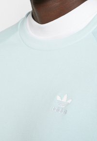 Ljusblå sweatshirt med vit krage, med en liten, broderad vit Adidas-logotyp på bröstet. Slät, bomullsliknande textur.