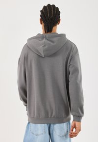 Felpa grigia con cappuccio, dal taglio rilassato, con polsini e orlo a coste. Presenta una tasca a marsupio e una texture liscia. Indossata con jeans azzurri chiari.