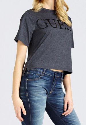 Kvinde iført mørkegrå cropped Guess t-shirt og blå jeans, står med hænder afslappet ved siderne, mod en enkel lys baggrund.