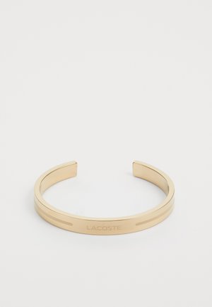 Bracelet jonc en métal doré avec texture lisse, présentant le marquage gravé "LACOSTE" et deux lignes parallèles le long du bandeau.