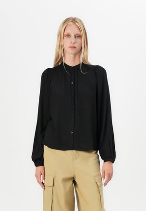 Schwarze Bluse mit langen Ärmeln, Knopfleiste, runder Ausschnitt und weichem Stoff. Verfügt über geraffte Schultern und entspannte Passform, kombiniert mit beige Farbhosen.