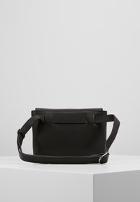 Černá kožená crossbody taška s texturou, nastavitelným popruhem a stříbrnou sponou, umístěná na bílém podstavci proti jednobarevnému šedému pozadí.