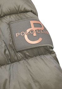 Olivgrön vadderad jacka med slät textur, med ett rektangulärt märke i mörkgrått med en kopparfärgad logga "POLO CLUB" och medföljande grafik.