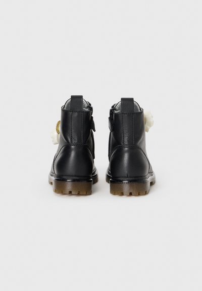 Par de botas de cuero negro hasta el tobillo con suelas marrones, vistas desde la parte trasera, cada una con una decoración de flor blanca en el lateral.