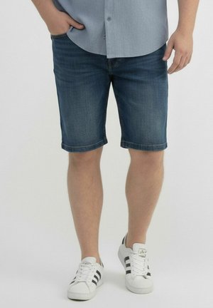 Mann trägt dunkelblaue Jeansshorts, hellblaues Hemd mit Knöpfen und weiße Turnschuhe mit schwarzen Streifen, steht mit einer Hand in der Tasche.