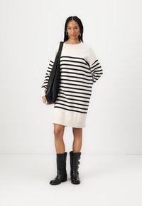 Sweater dress in cream with navy stripes, oversized design, ribbed hem, worn with knee-high black boots and a black shoulder bag.