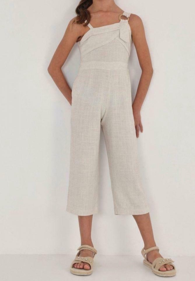 Femme portant une combinaison beige à une épaule avec des jambes larges coupées et des sandales beiges ornées de perles, debout sur un fond blanc.