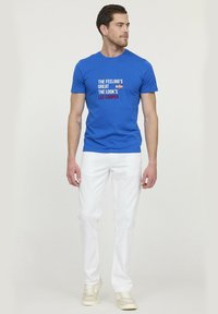 T-shirt en coton bleu avec un graphique textuel en blanc et rouge, associé à un pantalon blanc et des baskets légères, se tenant devant un fond uni.