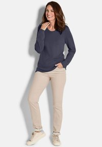 Lächelnde Frau, die mit einer Hand in der Tasche ihrer beigen Hose steht, trägt einen navy-farbenen Langarmpullover und beige Sneakers vor einem einfarbigen Hintergrund.