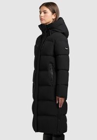 Dugi crni puffer kaput s kapuljačom, s patentnim zatvaračem na prsnom džepu, zatvaranjem na vezicu, quilted panelima i brendiranim logotipom na rukavu.