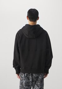 Černá mikina s kapucí a volným střihem, vybavená klokankovou kapsou a žebrovanými lemy na rukávech. Na zádech je našitý panel s detaily.