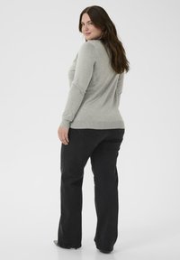 Vrouw met lang bruin haar, gekleed in een lichtgrijze lange mouwen top en zwarte jeans, staande en over haar schouder kijkend.