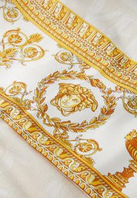 White silk scarf featuring intricate gold and yellow baroque patterns, including a central face, laurel leaves, and ornate accents.