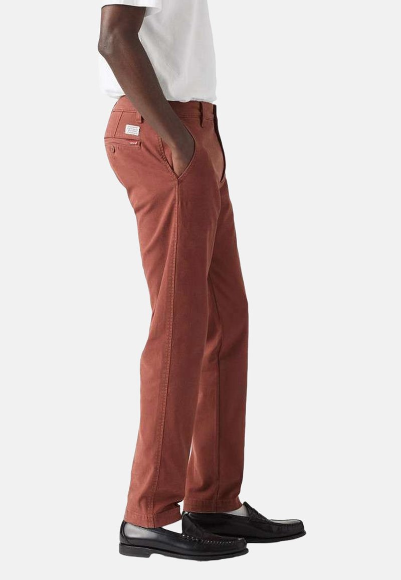 Rustfarvede bukser lavet af stof med en glat tekstur, der har sidelommer og syede detaljer, sammen med sorte sko.