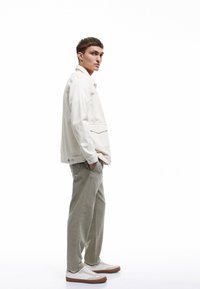 Lys cremefarvet denimjakke med en frontlomme, parret med lysegrønne bukser. Hvide sneakers med brune såler. Model poserer sidelæns.