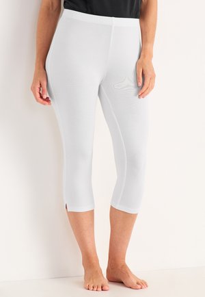 Kvinde iført tætsiddende hvide korte leggings med små sideslids, stående barfodet mod en lys baggrund.