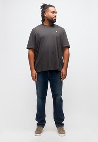 Άνδρας με ντρέντλοκς, φοράει σκούρο γκρι μπλουζάκι, μπλε τζιν και μπεζ αθλητικά παπούτσια, στέκεται και κοιτάζει προς τα πλάγια μπροστά σε απλό λευκό φόντο.