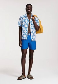 Camicia a maniche corte con motivo circolare blu e bianco, abbinata a pantaloncini da bagno blu tinta unita e sandali grigi. Borsa gialla a tracolla.