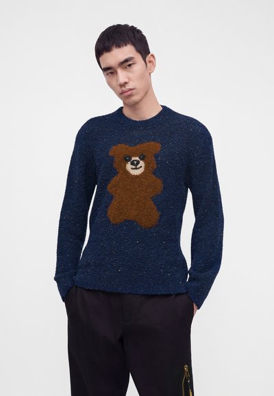 Jeune homme portant un pull texturé bleu marine avec un grand motif d'ours en peluche marron sur le devant, mains dans les poches d'un pantalon noir.