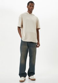 Huppari, jossa on kermaisen värinen ylisuurikokoinen malli ja tekstuuri-tasku. Yhdistetty löysiin sinisiin farkkuihin ja valkoisiin tennareihin, joissa on ruskeat korostukset.