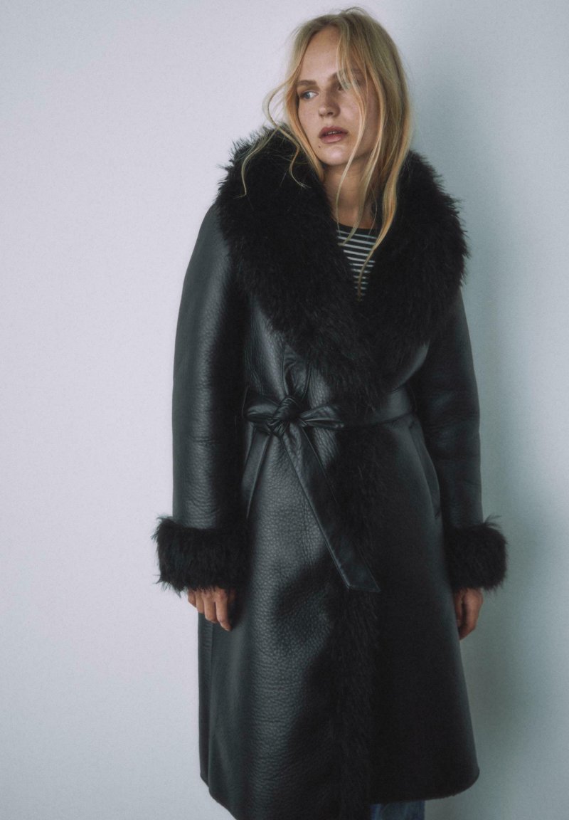 Czarny skórzany płaszcz z szerokim futrzanym kołnierzem i mankietami, zapinany paskiem w talii, o teksturowanym wykończeniu i długości do połowy łydek.