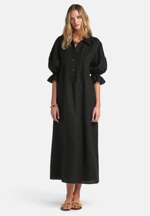 Žena u dugoj crnoj haljini s naboranim rukavima, kopčanom sprijeda, zlatnim nakitom i ispletenim ravnim sandalama, stoji na bijeloj pozadini.