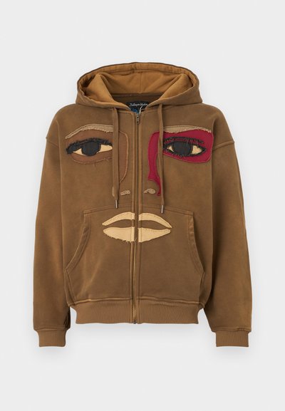 Brown zip-up hoodie made of soft fabric featuring a face design with stitched eyes and lips, highlighting contrast colors in red and beige.
