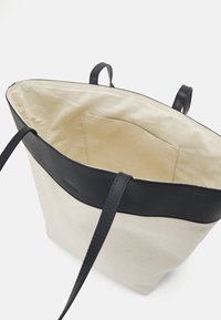 Sac fourre-tout en toile avec poignées et finitions en cuir noir, montrant un intérieur en tissu beige avec une poche intérieure.