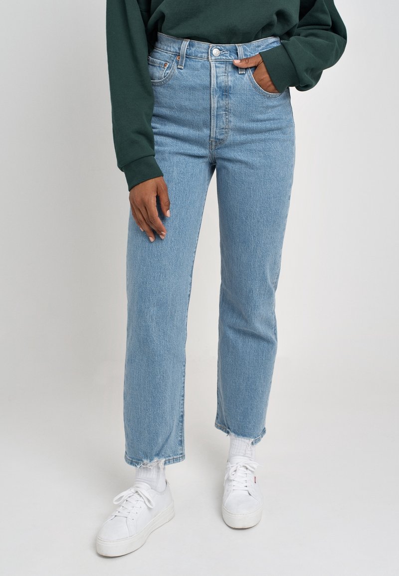 Jeans de talle alto en azul claro con pierna recta, bolsillos frontales y dobladillo deshecho, combinados con zapatillas blancas y un sudadera verde.
