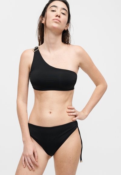 Γυναίκα φοράει μαύρο μπικίνι με έναν ώμο, με χρυσή αγκράφα και μαύρο κάτω μέρος μπικίνι με δεσίματα στο πλάι.