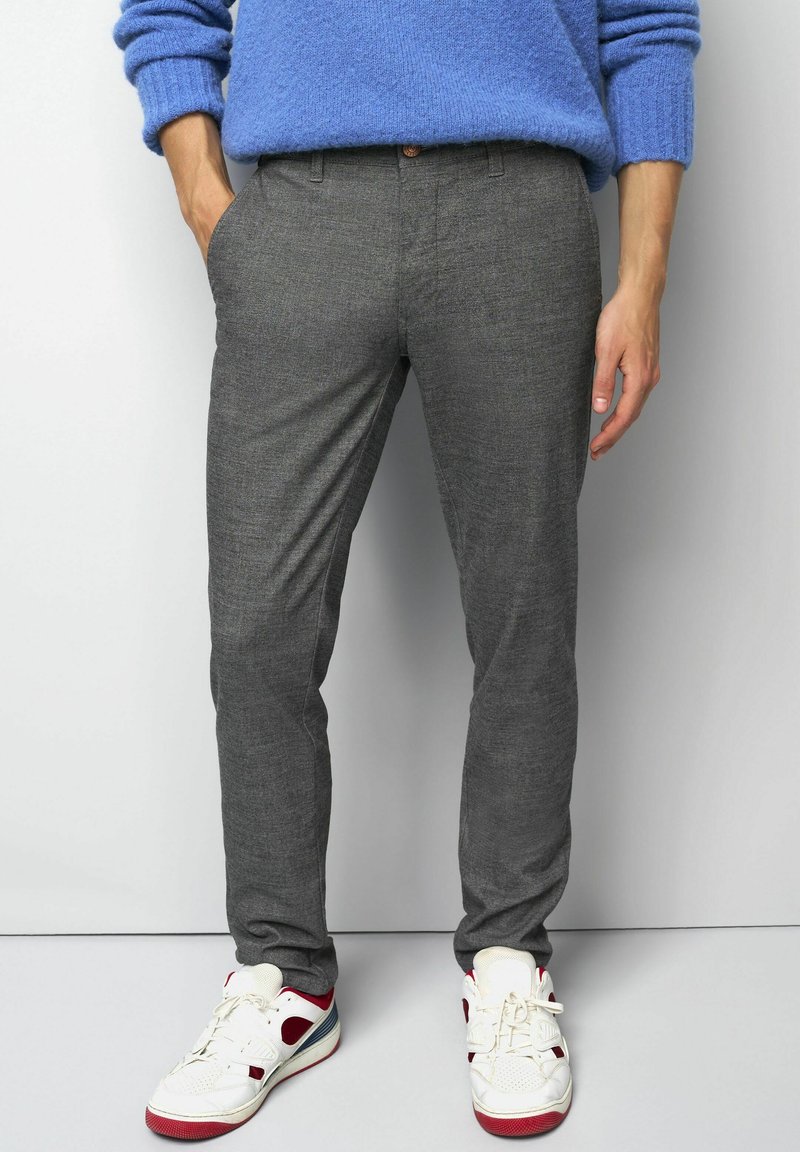 Mann, der enganliegende graue Hosen und weiße Sneakers mit roten und marineblauen Akzenten trägt, mit entspannten Händen an den Seiten vor einem schlichten Hintergrund.