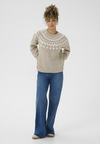 Pulover tricotat bej deschis cu un model geometric alb pe umeri, asociat cu blugi albaștri evazați și încălțăminte bej.