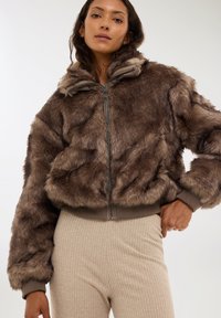 Giacca bomber in finta pelliccia color marrone con chiusura a zip, polsini e orlo a costine. Presenta una texture morbida e un cappuccio. Abbinata a pantaloni beige a costine.