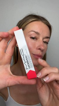 Donna che tiene una scatola di rossetto MAC Locked Kiss 24 ore vicino alla fotocamera con uno sfondo grigio neutro.