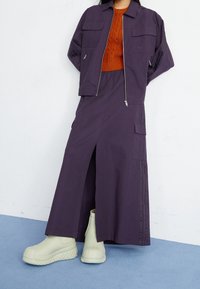 Lila kort jacka med två bröstfickor, i kombination med en matchande kjol med en slits. Chunkiga mintgröna gummistövlar fulländar outfiten.