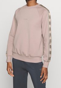 Sweatshirt rose clair avec des poignets et un ourlet côtelés. Il présente un col rond et un logo noir sur la poitrine, avec un accent à motif le long des manches.