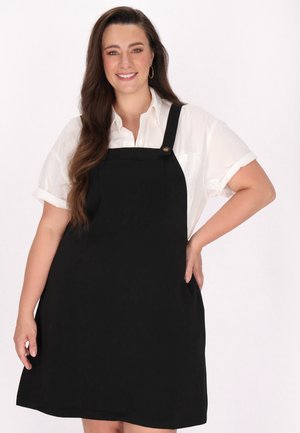 Rochie salopetă neagră cu bretele late și un buzunar frontal, purtată peste o cămașă albă cu nasturi; textură netedă și design până la genunchi.