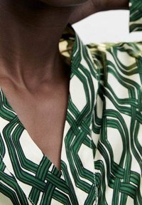 Groene en crèmekleurige blouse met een V-hals ontwerp, met geometrische lijnen en texturen, gemaakt van een lichtgewicht, gladde stof.