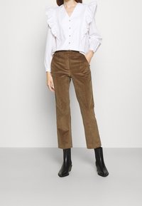 Witte blouse met ruffeldetails en knopen, gecombineerd met bruine corduroy broek. Zwarte enkellaarsjes maken de outfit compleet.