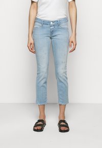 Personne portant un jean bleu clair courts déchiré, un t-shirt blanc et des sandales noires à bout ouvert, debout devant un fond blanc uni.