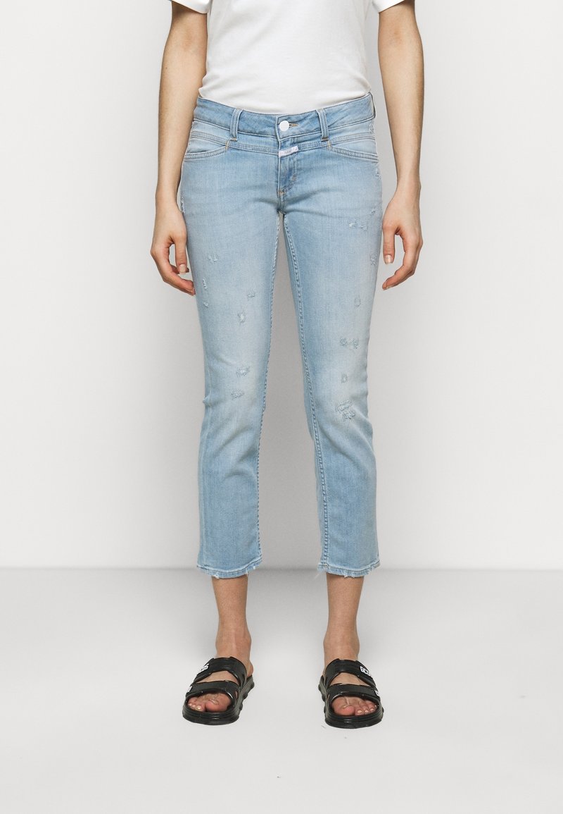 Personne portant un jean bleu clair courts déchiré, un t-shirt blanc et des sandales noires à bout ouvert, debout devant un fond blanc uni.