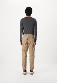 Beige cargo pants with elastic waistband, two side pockets, and ankle cuffs. Grey long-sleeve top visible. Neutral background.