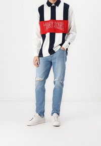 Long-sleeve polo shirt with navy and white stripes and a red horizontal band featuring "TOMMY JEANS." Paired with light blue ripped jeans and white sneakers.