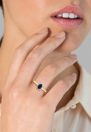 Guldring med en oval blå ædelsten omgivet af små klare sten, sat på en glat ring. Modellens hånd hviler nær hendes ansigt.