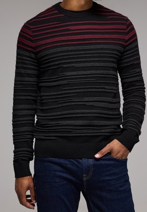 Mężczyzna w czarnym swetrze z teksturowanymi poziomymi paskami w ciemnoszarym i bordowym kolorze, w połączeniu z ciemnoniebieskimi dżinsami, z ręką w kieszeni.