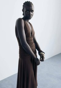 Robe sans manches marron avec un décolleté en V profond, détails froncés à la taille, jupe fluide, portée avec plusieurs bracelets aux poignets.