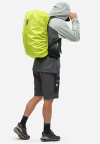 Jasnożółty wodoodporny plecak z zaciąganym zamknięciem, noszony na jasnoszarej i ciemnoszarej stylizacji z szortami i butami do trekkingu.